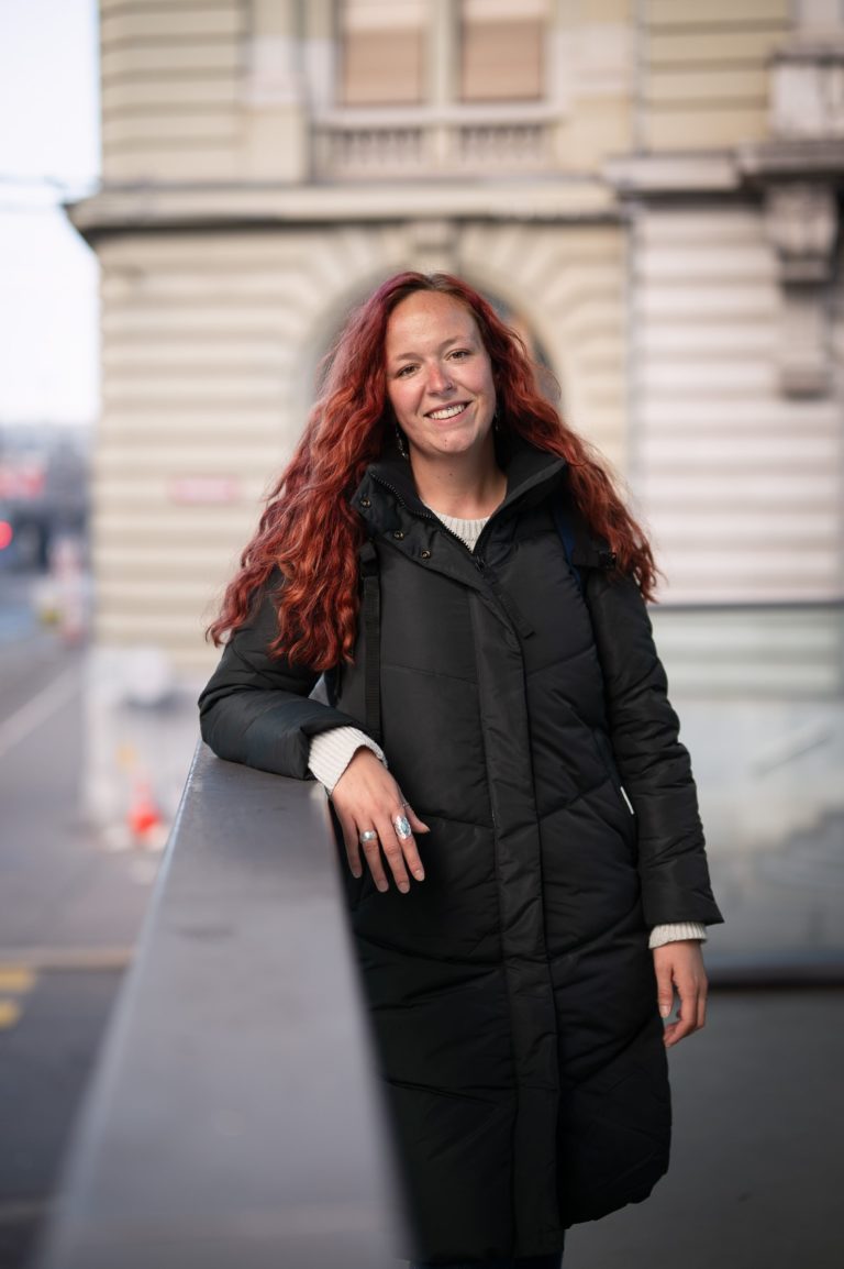 Sarina Portmann mit langen roten Haaren steht mit schwarzer Winterjacke auf einer Brücke in Bern und lacht direkt in die Kamera.