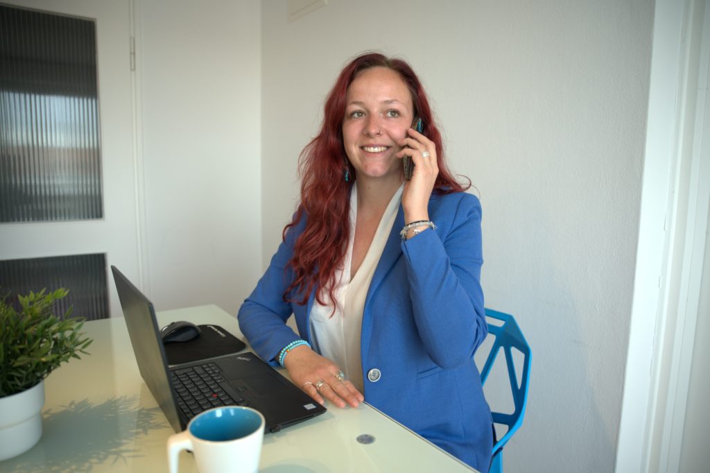 Sarina Portmann sitzt telefonierend auf einem blauen Stuhl am Tisch mit Laptop. Blauer Blazer & lange rote Haare trägt sie.
