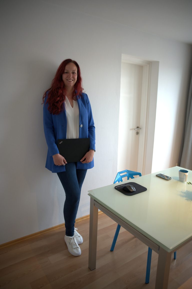 Sarina Portmann steht lächelnd hinter dem Tisch. Laptop in der Hand, Blauer Blazer & lange rote Haare sind zu sehen.