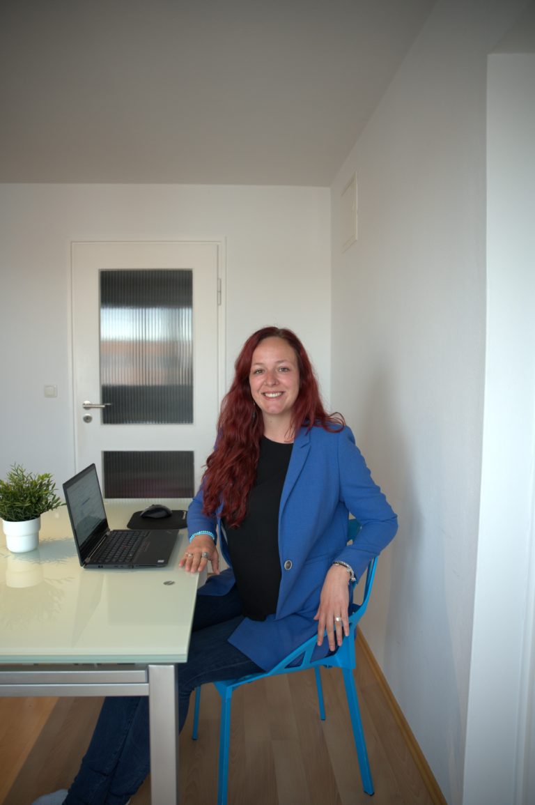 Sarina Portmann sitzt lächelnd auf einem blauen Stuhl am Tisch mit Laptop. Blauer Blazer & lange rote Haare trägt sie.