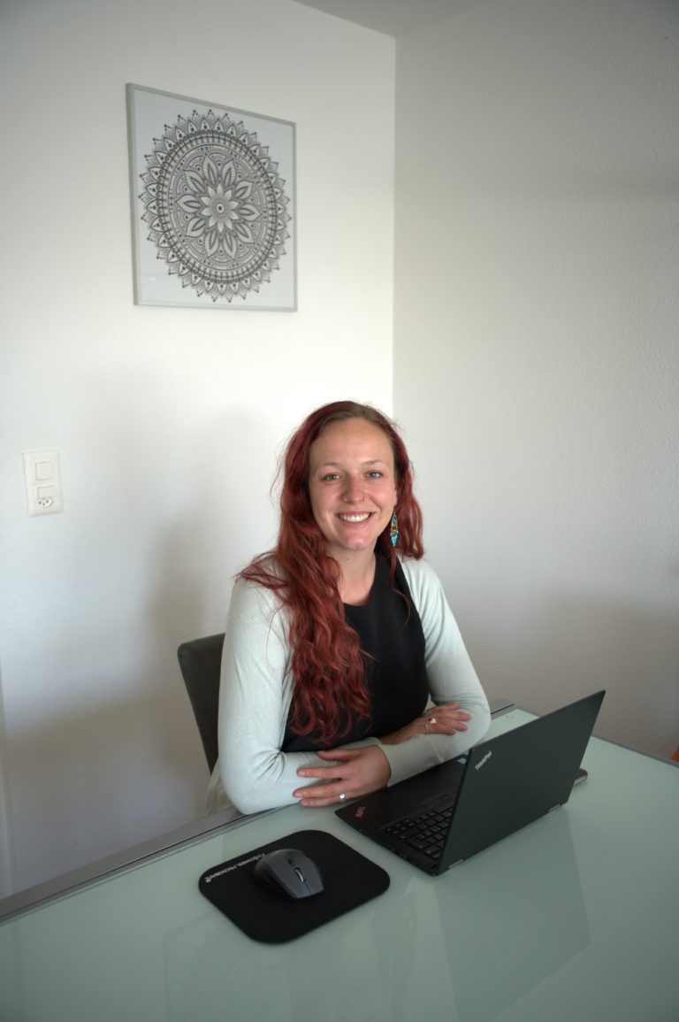 Sarina Portmann sitzt lächelnd auf einem Stuhl am Tisch mit Laptop mit langen roten Haare und einer hellblauen Jacke.