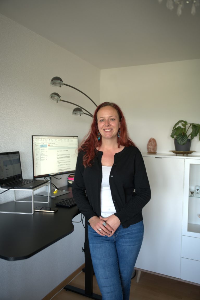 Sarina Portmann steht lächelnd vor dem Schreibtisch. Laptop, ein Bildschirm, eine Pflanze & lange rote Haare sind zu sehen.