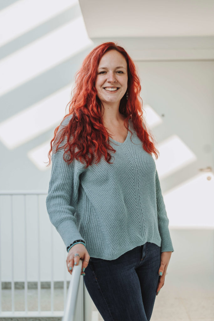 Sarina Portmann stehend an einem Treppengeländer mit weissem Hintergrund. Dunkelblaue Jeanshose, blauer Pullover, lange rote offene Haare und Sarina Portmann lacht direkt in die Kamera